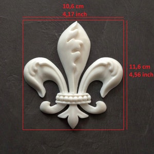 May include: A white fleur-de-lis decorative applique with a beaded band detail. The object measures 10.6 cm in width and 11.6 cm in height as indicated by the red measurement lines and text overlaid on the image.