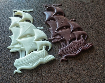 May include: Two decorative wooden ships, one white and one brown, are displayed on a brown textured surface. The ships are detailed with sails and a hull.