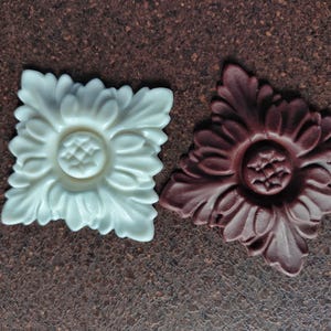 Könnte beinhalten: Zwei dekorative florale Verzierungen, eine weiße und eine braune, mit komplizierten Designs. Beide sind quadratisch und haben eine erhabene Mitte.