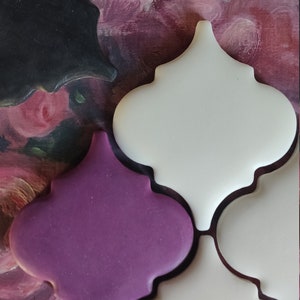 May include: Three ceramic tiles in a Moroccan style with a white, purple, and black glaze. The tiles are arranged in a pattern and are set against a background of pink and green floral painting.