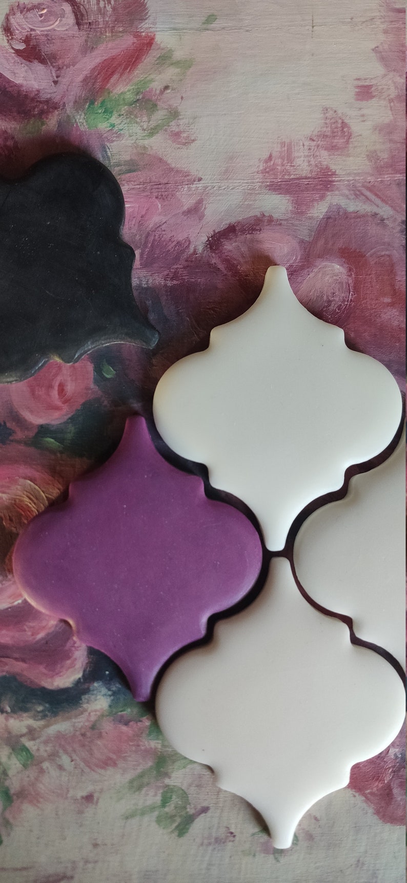 May include: Three ceramic tiles in a Moroccan style with a white, purple, and black glaze. The tiles are arranged in a pattern and are set against a background of pink and green floral painting.