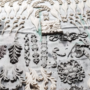 May include: A collection of white and gray decorative moldings, including floral, leaf, and scroll designs. The moldings are arranged on a white surface.