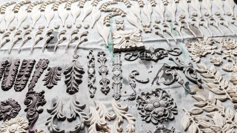 May include: A collection of white and gray decorative moldings, including floral, leaf, and scroll designs. The moldings are arranged on a white surface.
