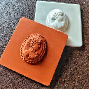 以下が含まれることがあります： カメオスタイルのジュエリーを作るためのシリコンモールド2個。左側のモールドは茶色で、右側のモールドは白。両方のモールドは、楕円形のフレームの中に女性の横顔のレリーフデザインが施されています。