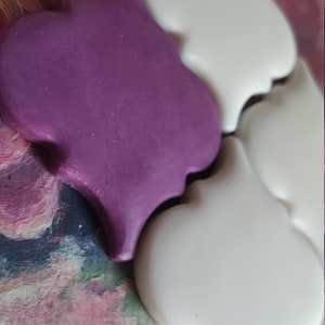 May include: Two ceramic tiles in the shape of a quatrefoil. One tile is purple and the other is white.