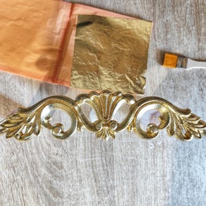 Puede incluir: Un tirador de mueble dorado y ornamentado con un diseño floral, junto a hojas de oro y un pequeño pincel. El tirador es un elemento decorativo, probablemente para un armario o cajón, y es de metal.