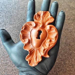 May include: A wooden heart-shaped keyhole with intricate carvings and a detailed design. The keyhole is a decorative element with a rustic, antique aesthetic.