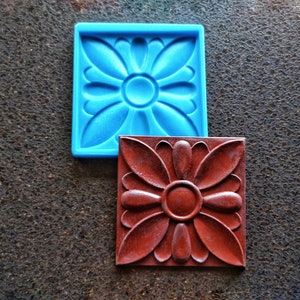 Könnte beinhalten: Zwei quadratische Silikonformen, eine blaue und eine braune, mit einem erhabenen Blumenmuster. Die Formen befinden sich auf einer strukturierten braunen Oberfläche.