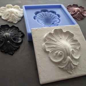 Op de afbeelding: Een siliconen mal voor het maken van decoratieve gips- of harsversieringen. De mal is blauw en heeft een enkele holte in de vorm van een gestileerd bloemmotief. Er zijn drie afgewerkte versieringen op de afbeelding, een witte, een zwarte en een bruine.