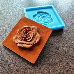 May include: Two silicone molds for making resin crafts. One mold is blue and features a raised rose design. The other mold is brown and features a raised rose design.
