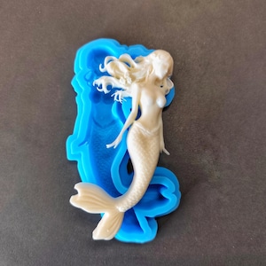 Pode incluir: Uma estatueta de sereia creme com cabelo esvoaçante e uma cauda detalhada, colocada dentro de um molde de silicone azul brilhante. A sereia é o ponto focal, mostrando elementos de design intrincados. O molde fornece um fundo contrastante.