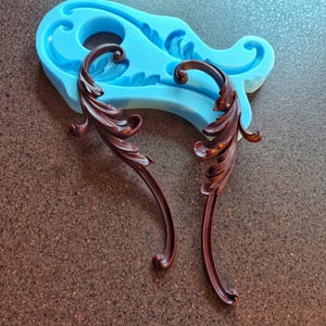 May include: Two dark brown, ornate, leaf-shaped decorative elements with a glossy finish. They are positioned near a light blue silicone mold with a matching leaf design. The items rest on a textured, brown surface.