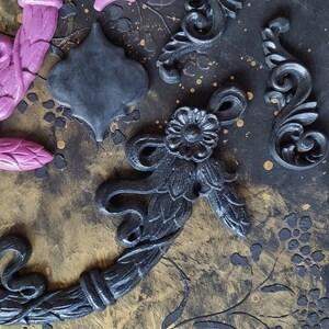 May include: A collection of decorative black and purple clay embellishments, including a wreath, a quatrefoil, and several ornate scrolls. The embellishments are arranged on a textured black and gold background.