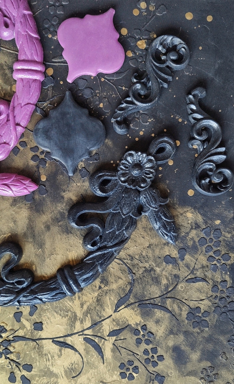 May include: A collection of decorative black and purple clay embellishments, including a wreath, a quatrefoil, and several ornate scrolls. The embellishments are arranged on a textured black and gold background.