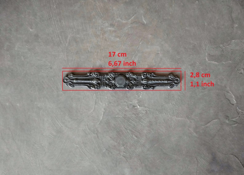 K&ouml;nnte beinhalten: Ein silberfarbener T&uuml;rgriff aus Metall mit filigranen Details. Er ist 17 cm lang und 2,8 cm breit.