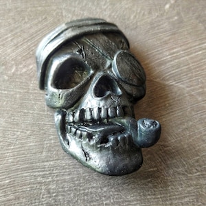 May include: A black skull figurine with a pirate eye patch and a pipe in its mouth. The skull is wearing a black bandana and has a detailed, textured surface.