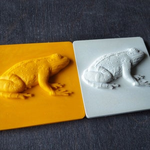 Könnte beinhalten: Zwei geformte Froschskulpturen aus Kunststoff, eine gelbe und eine weiße, auf einer dunklen Holzoberfläche.