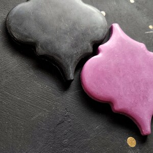 May include: Two decorative tiles in the shape of a quatrefoil. One tile is black and the other is a deep pink. The tiles are on a black background.