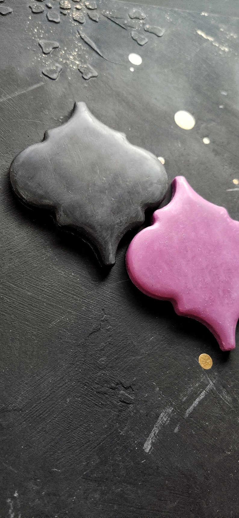May include: Two decorative tiles in the shape of a quatrefoil. One tile is black and the other is a deep pink. The tiles are on a black background.