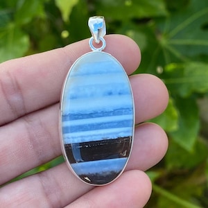 May include: Oval pendant with a silver-colored bail. The pendant features a layered design of blue and black bands. The stone is set in a silver-colored bezel. The background is blurred green foliage.