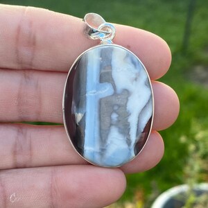 Peut inclure: Pendentif ovale avec une bélière argentée. Le pendentif présente une pierre polie avec un motif de tourbillons bleus, blancs et gris. La pierre est sertie dans un cadre en métal argenté. Le pendentif est tenu dans une main.