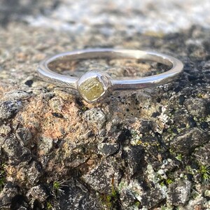 May include: A silver ring with a raw, light yellow diamond set in the center. The ring is displayed on a textured, brown and gray stone surface, with small patches of green moss.