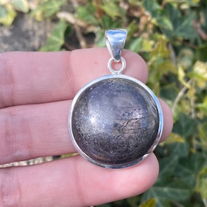 Puede incluir: Un colgante redondo de plata con una piedra oscura y moteada. La piedra tiene un brillo metálico y está engastada en un bisel de plata sencillo. El colgante tiene un pequeño lazo de plata para una cadena.