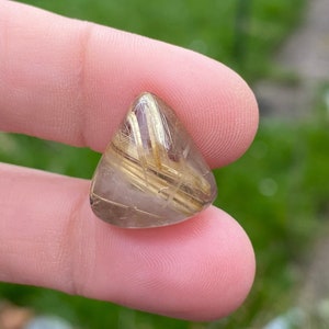 May include: A triangular, polished gemstone with golden, needle-like inclusions. The translucent stone has a light brown hue and is held between fingers against a blurred green background. The gemstone is approximately 2.5 cm in length.