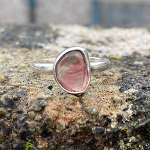 May include: A silver ring with an irregularly shaped gemstone in shades of pink and green. The stone is set in a bezel setting, and the band is smooth. The ring is shown on a textured stone surface. The ring is a piece of jewellery.