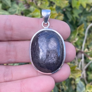 Peut inclure: Pendentif ovale avec une pierre sombre et réfléchissante sertie dans un cadre en métal argenté. La pierre a un aspect texturé et le pendentif comprend une boucle pour une chaîne. Le design est simple et élégant.