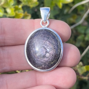 Puede incluir: Un colgante redondo con bisel y fianza plateados. El colgante presenta una piedra oscura e iridiscente con una superficie texturizada. La piedra tiene una mezcla de tonos morados, grises y plateados.