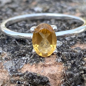 Peut inclure: Une bague en argent ornée d'une citrine ovale jaune doré. La bague a un fin anneau texturé, et la pierre est maintenue par quatre griffes. La bague est posée sur une surface de pierre texturée grise et brune.