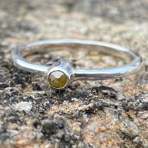 May include: A silver ring featuring a small, round, pale yellow gemstone at its centre. The ring is shown on a textured, grey and brown stone surface, with the band appearing smooth and polished.