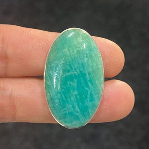 以下が含まれることがあります： 銀色のベゼルが付いた、楕円形のAmazonite宝石。石は明るいティール色で、自然な模様が見えます。宝石は、暗い背景に対して指の間に保持されています。