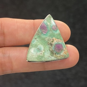 Op de afbeelding: Een driehoekige edelsteen met een zilveren rand. De steen heeft een gevlekte groene basis met onregelmatige vlekken van diep roze en rood. De edelsteen wordt tussen vingers gehouden tegen een donkere achtergrond.