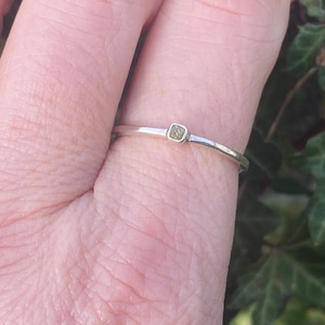 Puede incluir: Un delicado anillo de plata con una pequeña gema cuadrada de color verde pálido. El anillo tiene una banda fina y un diseño minimalista. La gema está engastada en un engaste sencillo. Una joya sutil para el uso diario.