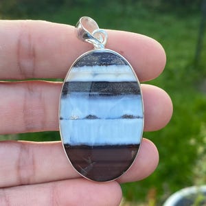 Peut inclure: Pendentif ovale avec une bélière en argent. Le pendentif présente un motif en couches de bandes marron, noires, blanches et bleu clair. La pierre est sertie dans une lunette en argent. Le pendentif est tenu dans une main.