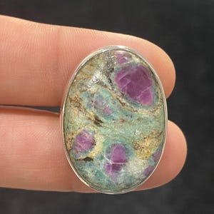 May include: Oval-shaped ring featuring a natural stone with a silver band. The stone has a mottled appearance with a mix of colors, including teal, tan, and vibrant magenta. The ring is a piece of jewelry.