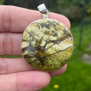 Peut inclure: Un pendentif rond avec une bélière argentée. Le pendentif présente une pierre polie avec un mélange de couleurs vertes, marron et beige. La pierre a un motif naturel unique. Le pendentif est probablement un bijou.