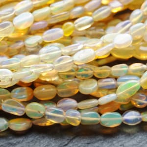 May include: Close-up of a collection of oval opal beads. The beads are translucent and iridescent, with colours from white and yellow to green and blue. The beads are strung together, suggesting they are for jewellery making.