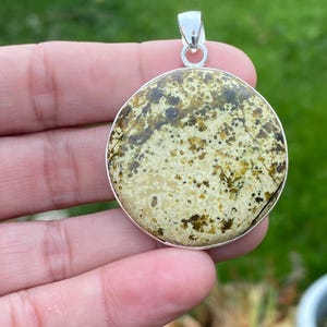 Peut inclure: Un pendentif rond avec une bélière argentée. Le pendentif présente un motif tacheté de tons vert clair, marron et beige. Le pendentif est tenu dans une main sur un fond vert flou.