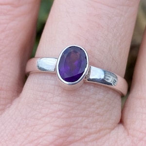 Könnte beinhalten: Ein Silberring mit einem leuchtenden, oval geschliffenen, violetten Edelstein. Der Edelstein ist in einer Zargenfassung gefasst, und das Band ist aus poliertem Silber. Der Ring wird an einem Finger gezeigt.