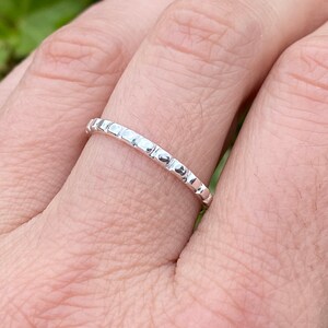 May include: A silver ring with a textured, dotted design. The ring is worn on a finger, showcasing its delicate and detailed craftsmanship. The ring's surface reflects light, highlighting its polished finish. A piece of jewelry.