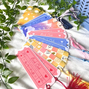 May include: A set of six colorful bookmarks with a book club theme. Each bookmark features a different design with a star, clouds, checkered pattern, and a rainbow. The bookmarks have a ticket shape and include the text "Book Club" and "Once upon a time".