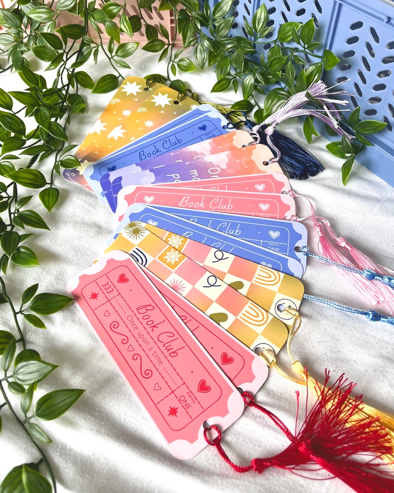 May include: A set of six colorful bookmarks with a book club theme. Each bookmark features a different design with a star, clouds, checkered pattern, and a rainbow. The bookmarks have a ticket shape and include the text "Book Club" and "Once upon a time".