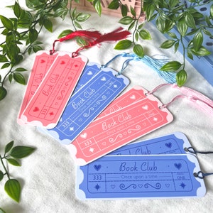 May include: A set of six bookmarks with a ticket design. The bookmarks are pink and blue with white text that reads "Book Club - Once upon a time". Each bookmark has a star, hearts, and a number 333 printed on it. The bookmarks have tassels in pink, blue, and red.