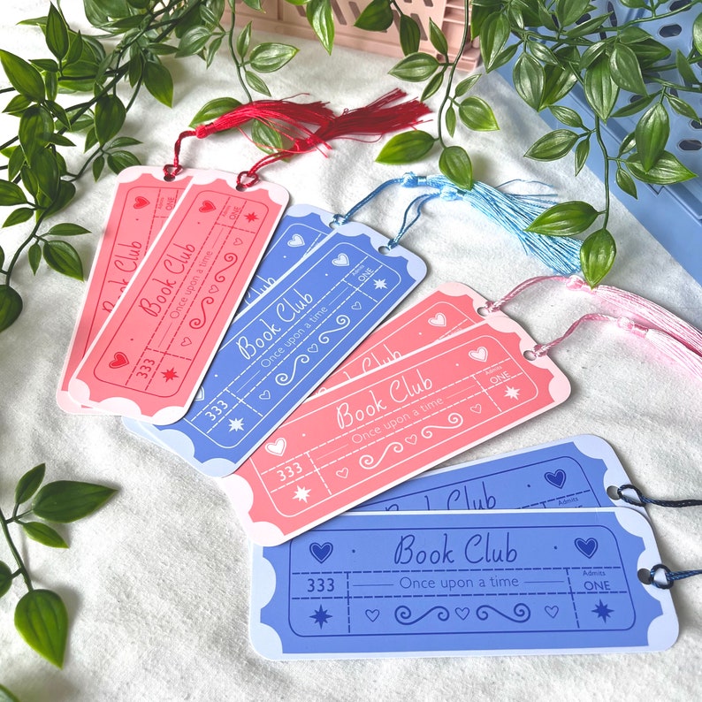 May include: A set of six bookmarks with a ticket design. The bookmarks are pink and blue with white text that reads "Book Club - Once upon a time". Each bookmark has a star, hearts, and a number 333 printed on it. The bookmarks have tassels in pink, blue, and red.