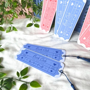May include: Two blue and two pink bookmarks with a ticket design. The bookmarks have the text "Book Club" and "Once upon a time" on them. The bookmarks have a star, hearts, and the word "ONE" on them. The bookmarks have a tassel.