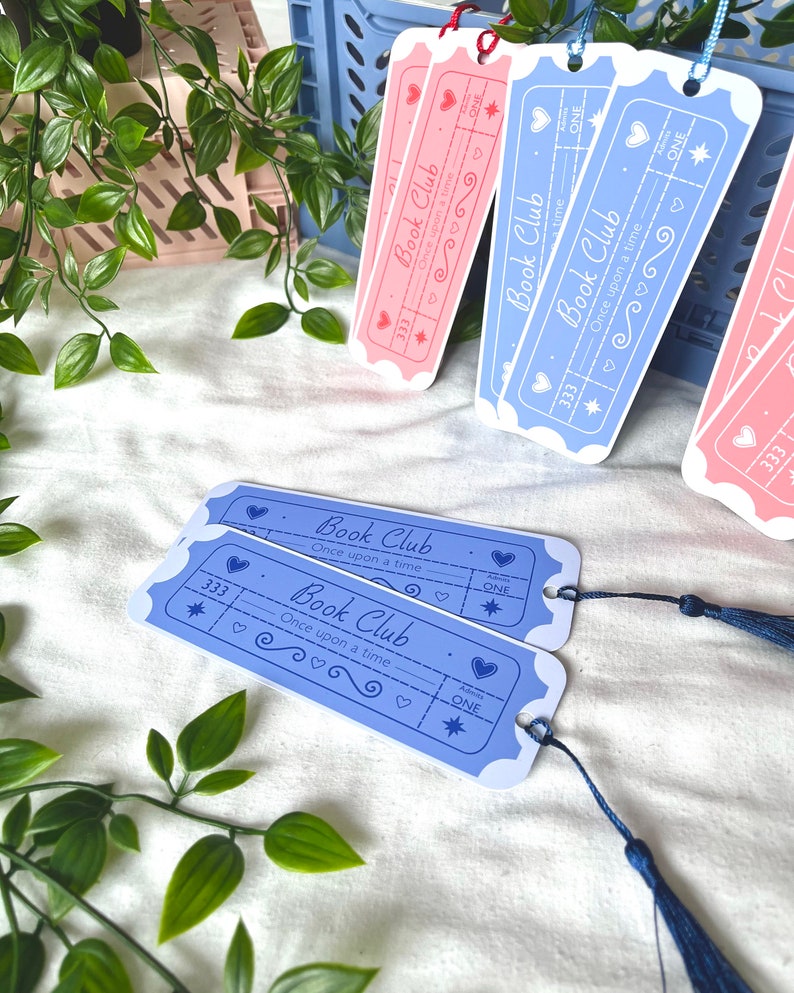 May include: Two blue and two pink bookmarks with a ticket design. The bookmarks have the text "Book Club" and "Once upon a time" on them. The bookmarks have a star, hearts, and the word "ONE" on them. The bookmarks have a tassel.