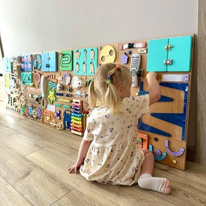 Könnte beinhalten: Ein Kind spielt mit einem Holz-Aktivitätsbrett mit verschiedenen Texturen, Farben und Formen. Das Brett verfügt über verschiedene interaktive Elemente, darunter ein Schloss, einen Spiegel, ein Telefon, ein Zahnrad, ein Labyrinth und ein Xylophon. Das Brett ist an einer Wand montiert und das Kind sitzt auf dem Boden davor.
